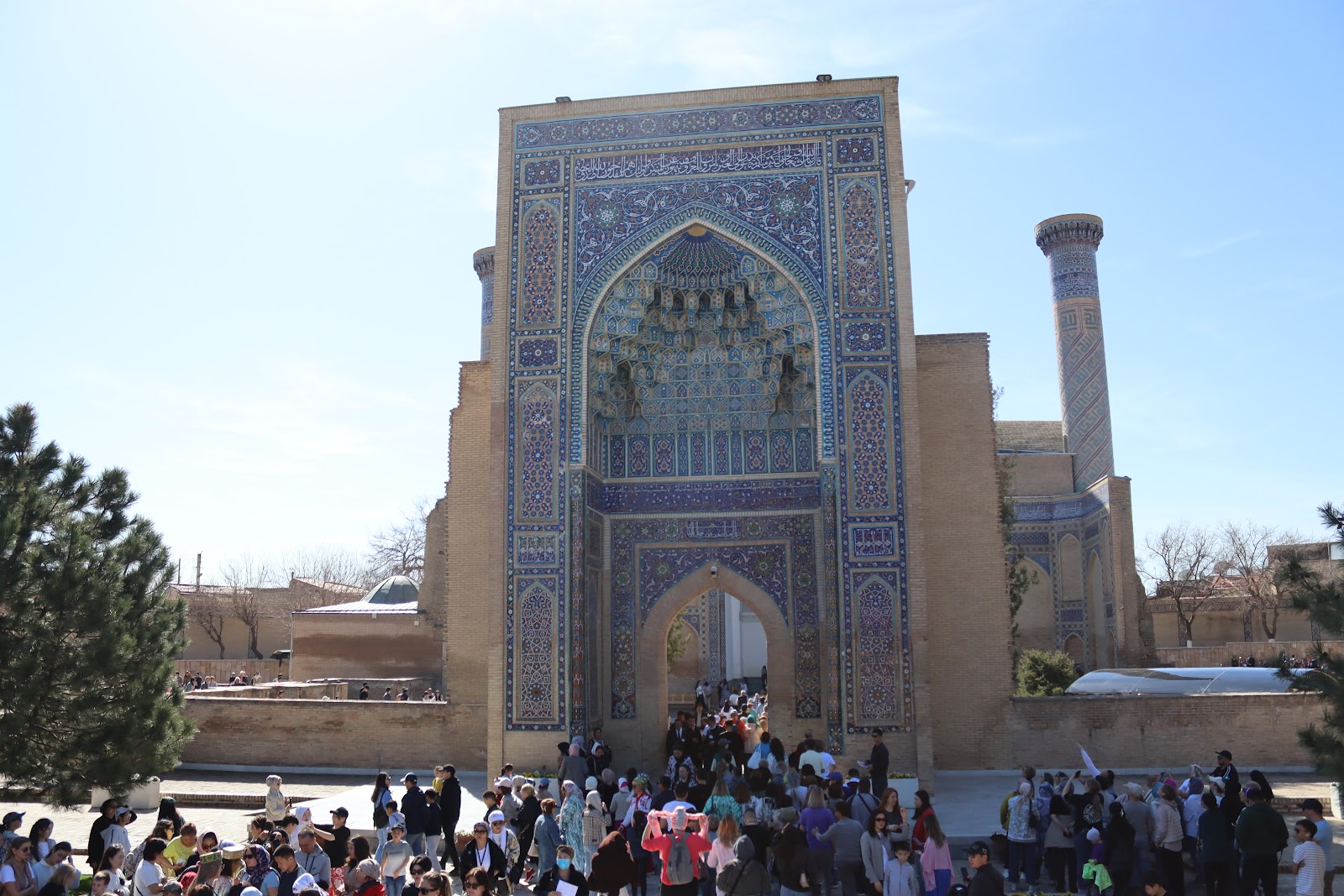 Ornate blue-tiled entrance portal of the Gur-e-Amir mausoleum crowded with visitors on a clear spring day
