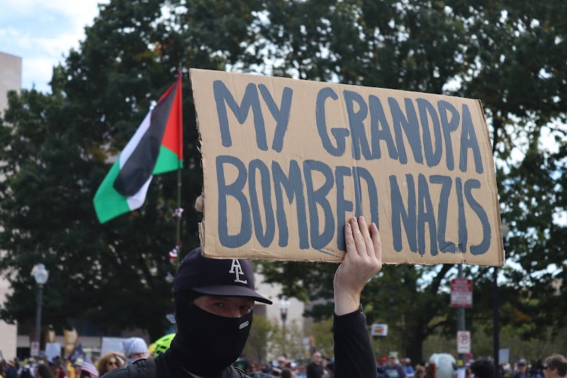 Protester holding a cardboard sign reading 'My Grandpa Bombed Nazis' at a march in Washington D.C., Palestinian flag visible behind