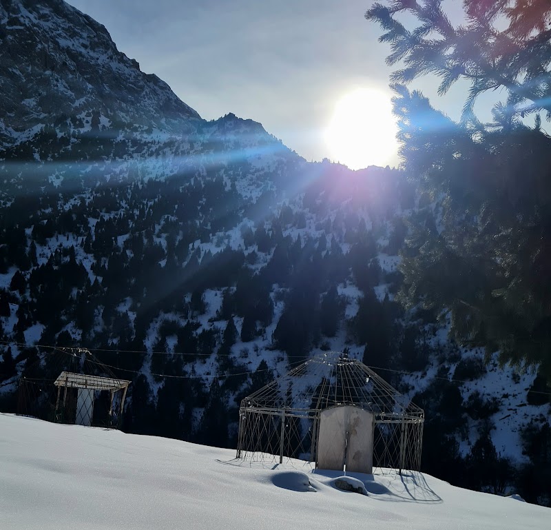 Skeletal yurt frames in deep snow against a forested mountain with a halo of winter sun