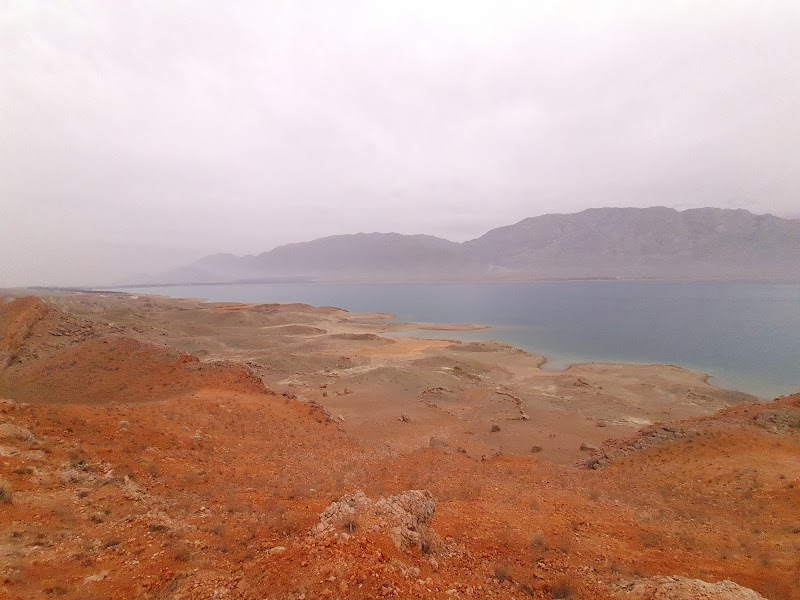 Rust-red rocky shoreline overlooking Toktogul Reservoir under an overcast sky, with mountains fading into the distance