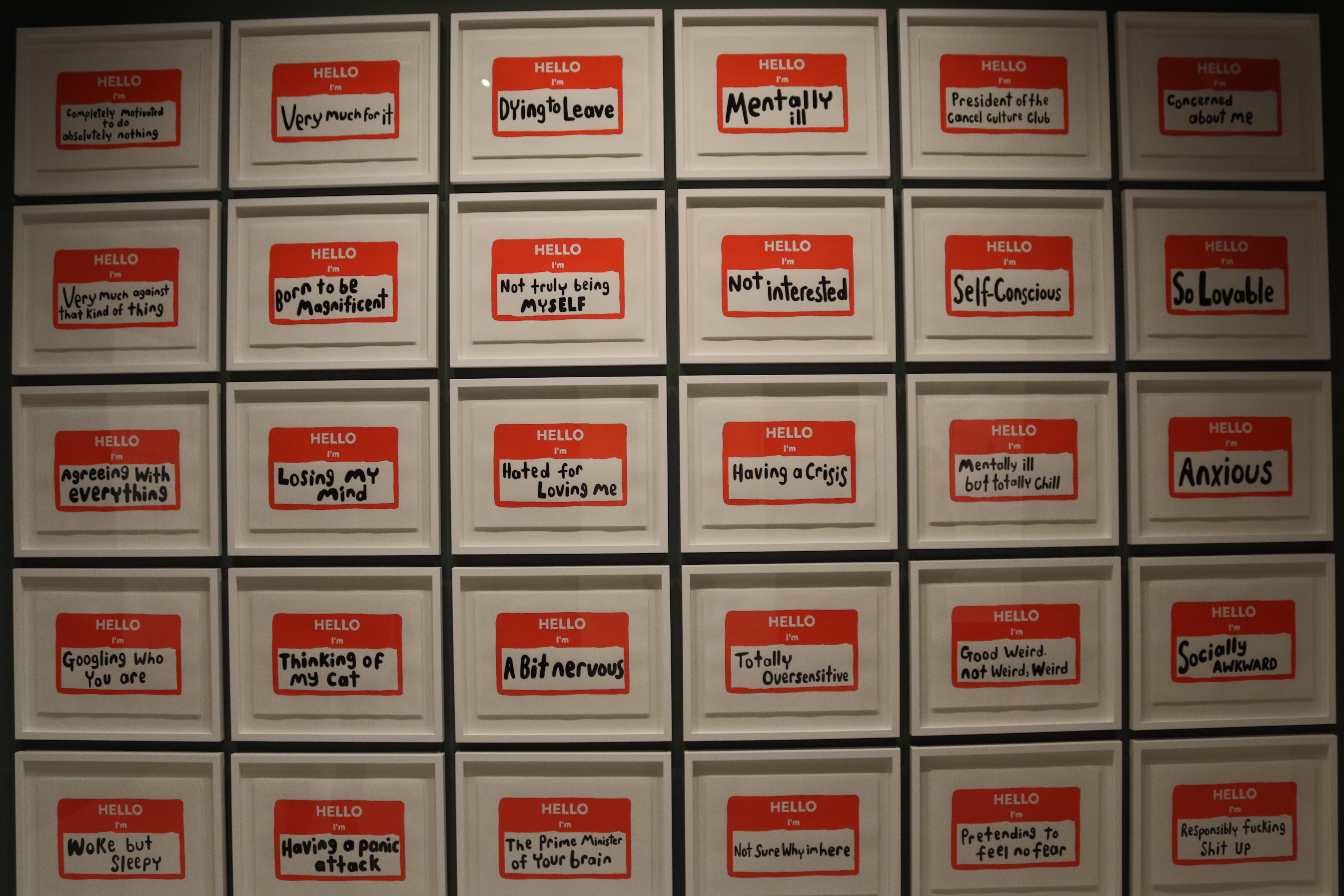 Grid of framed red HELLO name-tag artworks bearing humorous and anxious self-descriptions on a gallery wall