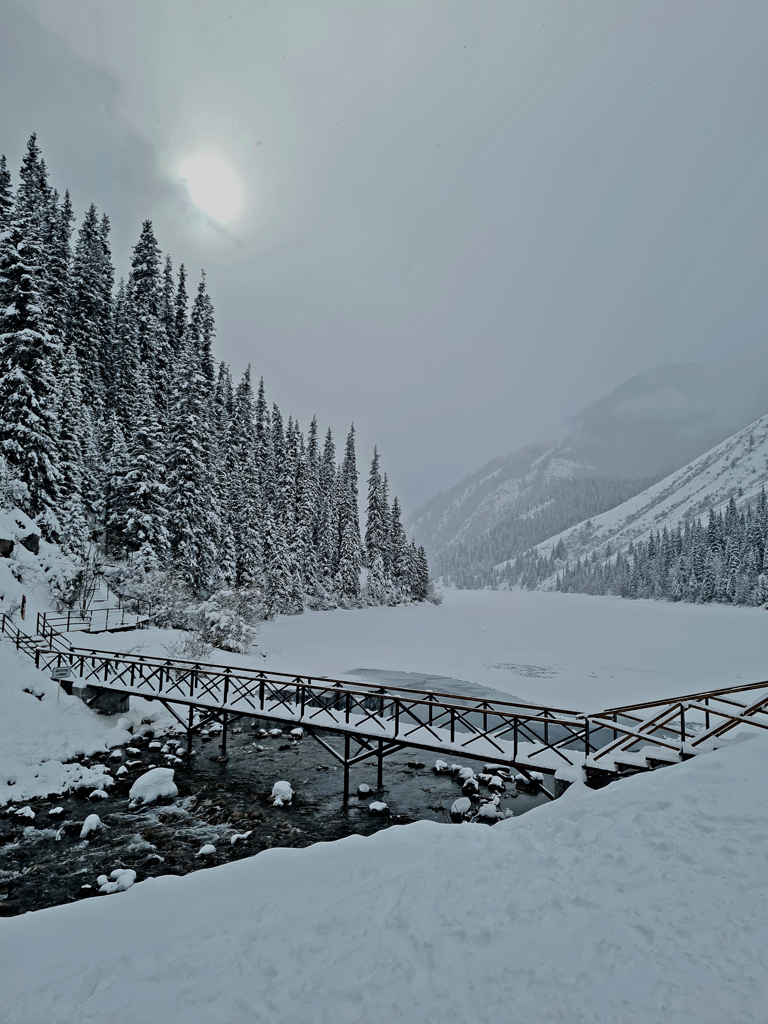 Wooden footbridge over a rushing stream leading to a snow-blanketed frozen lake ringed by snow-laden spruce trees
