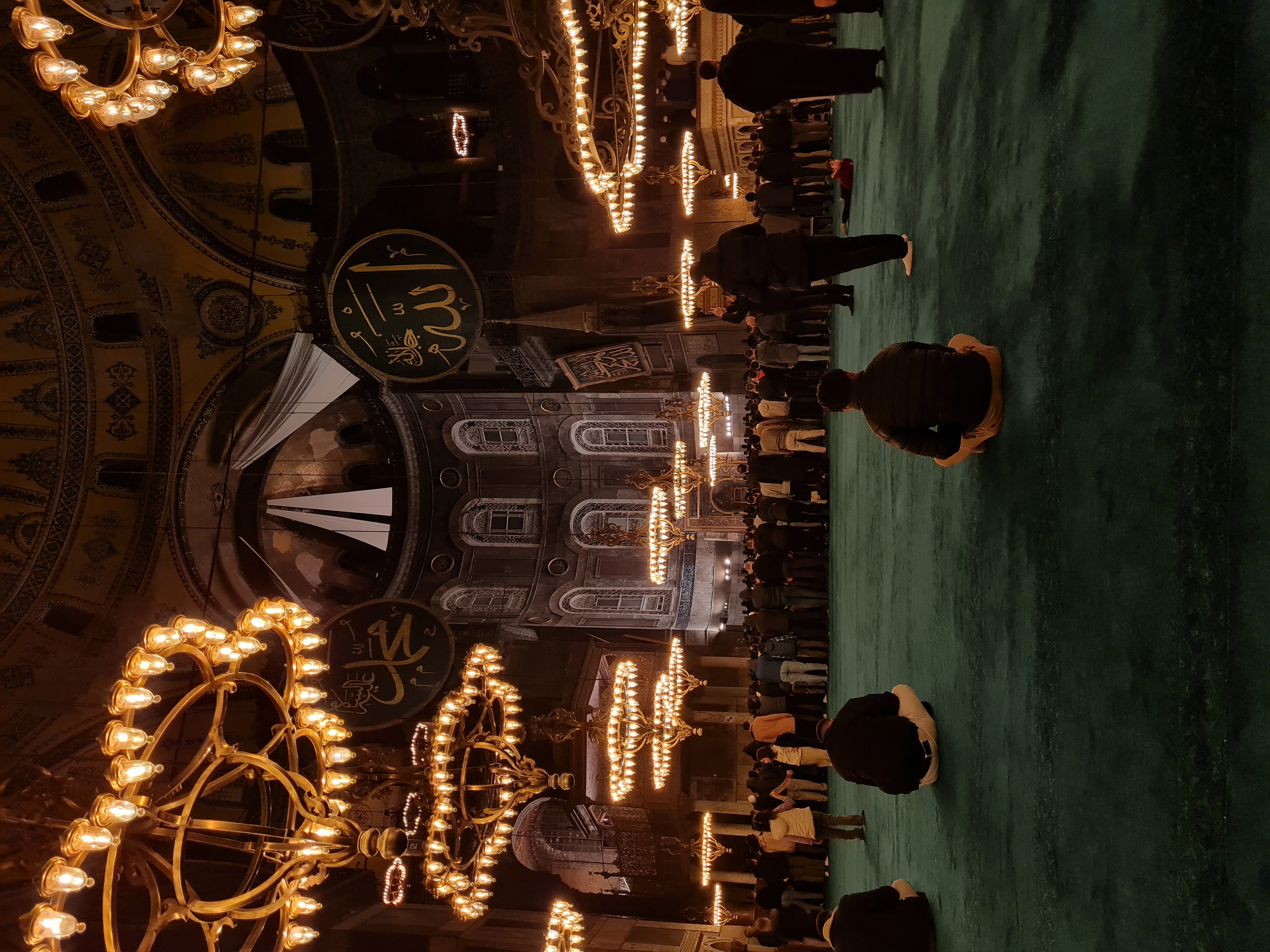 Interior of Hagia Sophia at night, golden chandeliers glowing against the vast dome with visitors silhouetted below