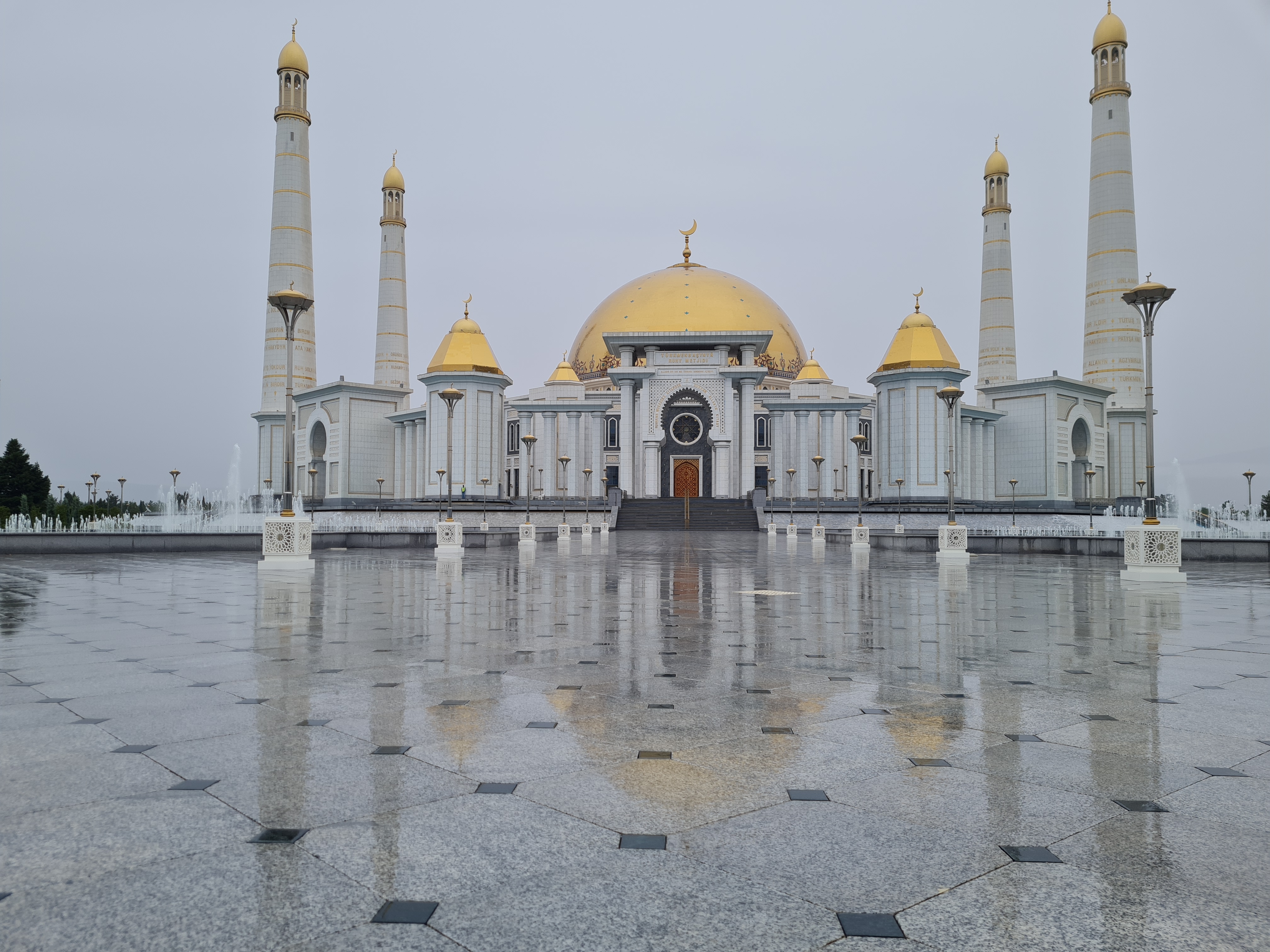 Vast white marble Ruhy Mosque with gold domes and four slender minarets perfectly reflected in a rain-soaked plaza
