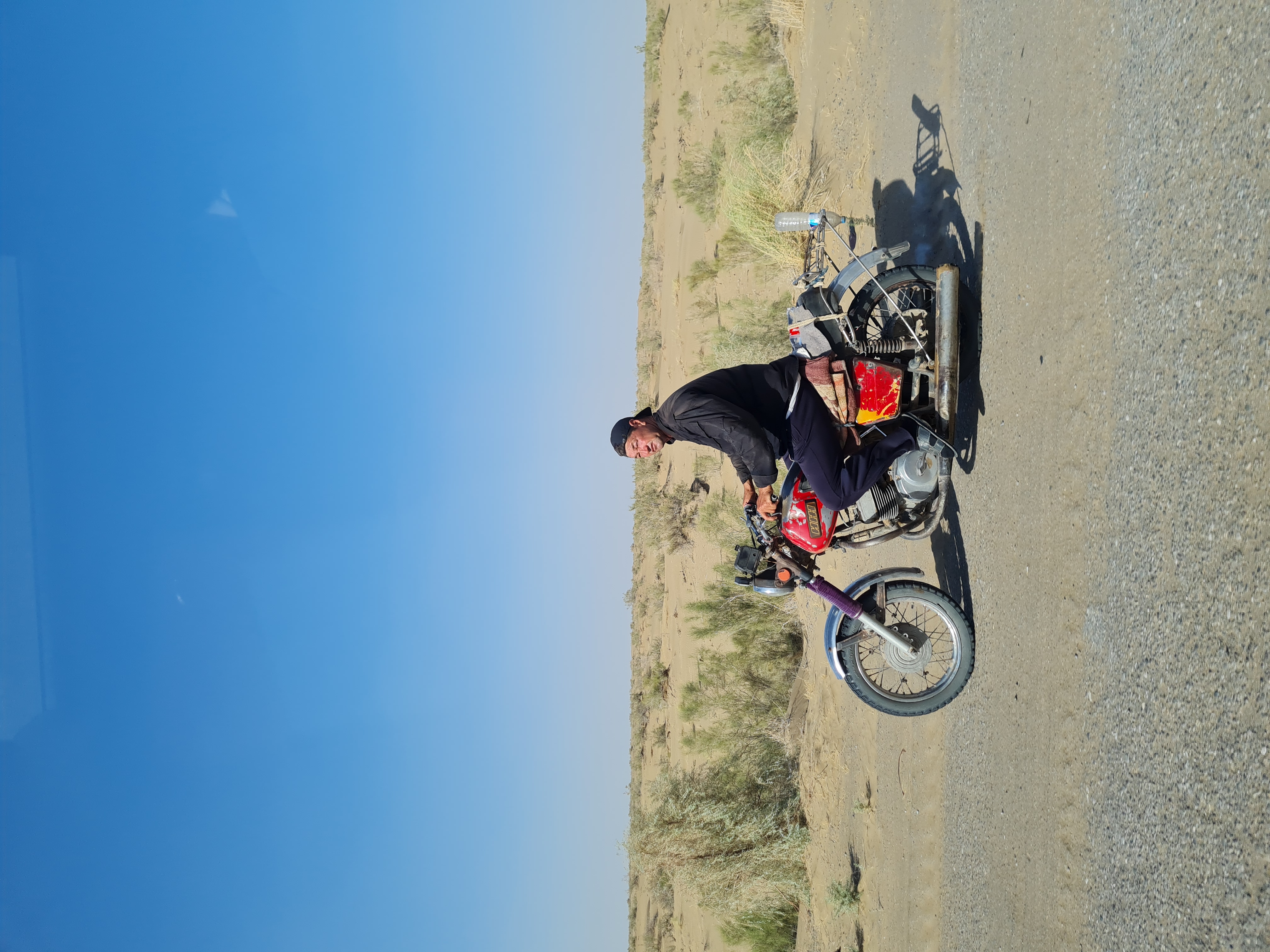 Man astride a red motorcycle on the flat desert highway leading to the Darvaza gas crater under a cloudless sky