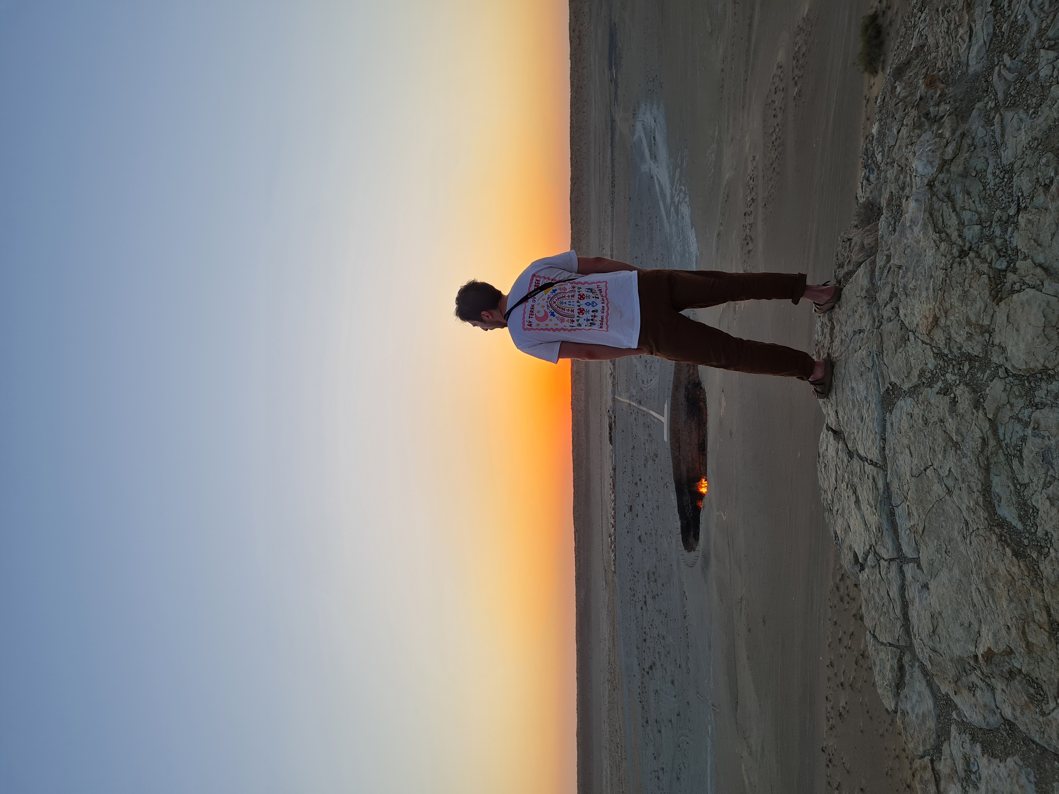 Silhouette of a person standing at the edge of the Darvaza gas crater, its orange flame glowing against a desert sunset