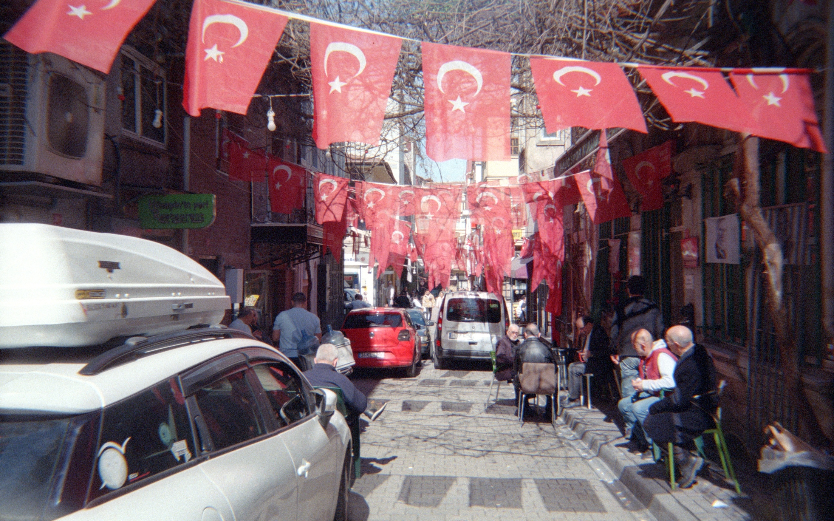 Narrow old-city lane draped end-to-end with red Turkish crescent flags, locals seated at café tables below